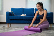 © PaeGAG - Asian sportswoman preparing for home workout, rolling a purple yoga mat in living room