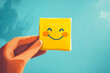 © Fleuronica - A hand presents a vibrant yellow sticky note adorned with a smiling face, set against a serene blue backdrop, exuding positivity