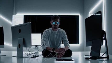 Young programmer wearing futuristic glasses sits on the floor in a modern office, surrounded by computer screens, looking directly at the camera