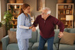 © Stockphotodirectors - A female home care nurse in scrubs assists an elderly man using a cane to stand up from his couch. The nurse holds his arm while the man uses his cane for support.