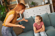© Stockphotodirectors - In a warm living room, a mother expresses her anger towards her daughter, who sits quietly with her arms crossed, conveying a tense exchange of emotions in their home environment.