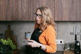 Blond woman in her kitchen prepares espresso using a capsule coffee machine and enjoys her coffee, embracing a cozy lifestyle.