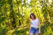 © SergeiPh - A beautiful woman stands in the forest on a summer day. She is wearing a white shirt and blue shorts. The scene is peaceful and serene as the woman is surrounded by nature