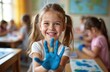 © Viktor - Smiling girl shows blue painted hand in classroom. Child looks at camera with cheerful expression. School lesson, art class, creative learning with classmates. Positive emotions, childhood happiness.