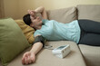 © Yuliia - woman lies on couch with arm extended, measuring blood pressure at home, showing tired or unwell expression
