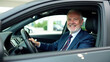 © yana136 - elderly man with a beard and glasses drives a modern car, wearing a suit and a white shirt, looking focused. background blurred city street and parked cars, transportation, senior lifestyle, business.