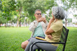 © bongkarn - Glasses old man holding smartphone taking picture of a woman while sitting together on bench in park