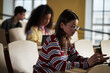 © DragonImages - Caucasian young woman sitting in lecture hall using digital tablet, multiethnic university students studying in background, focused on academic work, casual classroom setting