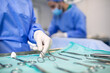 © Graphicroyalty - Close-up of a gloved hand selecting a surgical instrument from a sterile tray. In the background, medical staff prepare for the procedure in the operating room.