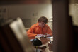 © TrueFrame Collective - Young adult Caucasian man sitting at table reading book in prison library, focused expression, short curly hair, surrounded by bookshelves and study materials in institutional setting