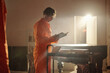 © TrueFrame Collective - Young adult Caucasian man standing in prison library reading book while holding cart with stacked books, focused on pages, wearing orange jumpsuit, illuminated by bright light