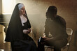 © TrueFrame Collective - Caucasian nun sitting beside Black priest holding book, both engaged in conversation in prison library setting, sunlight streaming through window in background