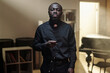 © TrueFrame Collective - Portrait of Black priest standing in prison library holding rosary beads, looking directly at camera with serious expression, bookshelves and piano in background