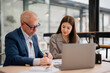 © MINAE - Senior businessman and young Asian businesswoman collaborating on a project, analyzing financial charts and data on a laptop in a modern office environment