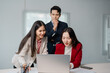 © Tj - Two asian businesswomen working together using laptop in office, analyzing financial statistics displayed on the screen, while a businessman standing behind them observes their work