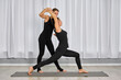 © TRAVELARIUM - Two adult women practice yoga in serene studio. Instructor assists student in warrior pose. Bright lighting and minimalist design create calmness
