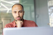 © Liubomir - A pensive, dark-skinned man with a beard rests his chin on his hand in front of a laptop, lost in thought.