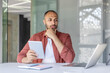 © Liubomir - A thoughtful man at a desk holds a tablet, pondering ideas while working on a laptop in a modern office setting.