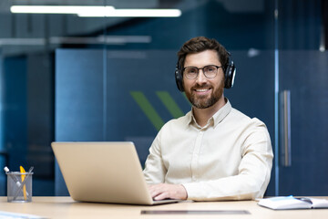 Portrait of a young man wearing headphones sitting at a desk in the office, working on a laptop and looking at the camera with a smile