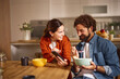 © Stockphotodirectors - A couple sits at a wooden table in a home office, smiling and engaging with each other while enjoying their breakfast from colorful bowls. Laughter fills the space.