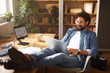 © Stockphotodirectors - In a well-lit home office, a man sits comfortably in front of a laptop, feet up on a desk, enjoying a relaxed work environment filled with plants and books on shelves.