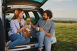 © Stockphotodirectors - Two friends are seated at the back of their car enjoying a cozy picnic in a beautiful outdoor location. They are sharing warm drinks while chatting happily in the golden light.