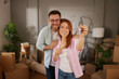 © Stockphotodirectors - A couple happily shows off their new keys in a cozy living room filled with unpacked boxes and personal touches, symbolizing the start of their journey in a new home.