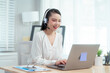 © Thitaporn - Asian woman wearing headset works at her desk in home office, engaging in online customer service and technical support, professional communication and leadership during virtual meetings and webinars