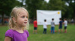 © Vasiliy - Little blonde girl in purple shirt looking thoughtfully at outdoor school event with presentation board. Child for educational activities and learning programs