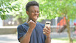 © stockbakers - Casual African Man Celebrating with Phone Outdoors