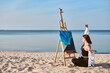 © TRAVELARIUM - Young woman paints abstract ocean scene on canvas, seated on sandy beach. Tranquil sea setting enhances creativity, symbolizing inspiration and serenity