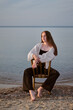© TRAVELARIUM - Young woman in casual attire sits on chair at serene beach, embodying calmness and introspection. Soft evening light and water create peaceful, reflective mood