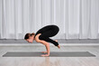 © TRAVELARIUM - Young, caucasian woman in black outfit performs crow pose on mat in minimalist studio with white curtains. Scene exudes focus and calm, highlighting balance and strength