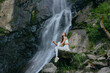 © SHOTPRIME STUDIO - Woman meditating near waterfall dressed in white activewear sitting peacefully on rocks surrounded by lush greenery and natural scenery in outdoor environment.