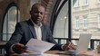 © Yuliia - African American entrepreneur typing laptop computer check data in papers dissatisfied problem mistake report bad result sad stress business man office businessman male guy research analyse documents