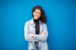 © mimagephotos - Confident woman with curly hair standing against blue wall, smiling with arms crossed in a casual and relaxed pose.