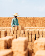 © Kowit - A man wearing a straw hat stacks rectangular bricks in an outdoor setting with large piles of similar bricks in the background under a clear blue sky.