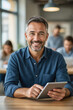 © f_bossa - A smiling middle-aged man in a blue shirt holds a tablet while seated in a modern office setting.