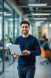 © f_bossa - A young man with dark hair and a beard confidently smiles while holding a laptop in a modern office setting.