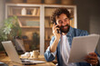 © Stockphotodirectors - A man with a beard holds papers in one hand and talks on the phone with a smile. He sits at a wooden desk, surrounded by books and plants, in a well-lit home office.