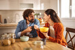 © Stockphotodirectors - A couple sits at a wooden table in a well-lit home office, sharing a light breakfast with smiles, enjoying each other's company and a moment of connection.