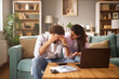 © Stockphotodirectors - A man is visibly stressed, holding his head in his hands, while a woman seated next to him points to financial documents on a laptop, both confronting their financial issues.