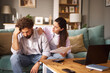© Stockphotodirectors - A couple sits on a couch in their living room, discussing financial difficulties. The woman comforts the man, who appears worried, holding documents while a laptop lies on the table.