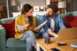 © Stockphotodirectors - A couple engages in a serious discussion about their financial issues while sitting together on a couch, surrounded by documents, a laptop, and a coffee mug at their home.