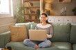 © Stockphotodirectors - Smiling woman with headphones sits on a green sofa, engaged in a virtual meeting on her laptop. The room is filled with books and warm lighting, creating a relaxed atmosphere.