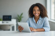 © f_bossa - A smiling Black woman in a light blue shirt holds a tablet while seated at a desk in a modern office setting.