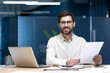 © Tetiana - Portrait of a smiling and successful young businessman sitting at a desk in an office center, working on a laptop and with documents