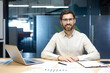 © Tetiana - Portrait of a young smiling man in a shirt and glasses sitting in the office at a table with a laptop and documents, looking at the camera