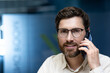 © Tetiana - Close-up portrait of a young male businessman in the office, talking on the phone and looking at the camera with a smile
