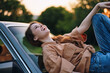 © SHOTPRIME STUDIO - Young woman laughing joyfully on a car hood during sunset, wearing a casual brown shirt and blue jeans, showcasing a carefree lifestyle in nature.
